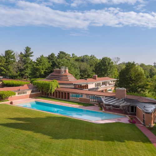 A large, uniquely designed house with a swimming pool, surrounded by green lawns and trees under a clear blue sky.