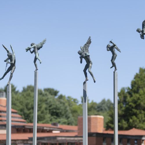 Sculptures of winged figures on tall poles, set against a background of trees and a building, under a clear blue sky.