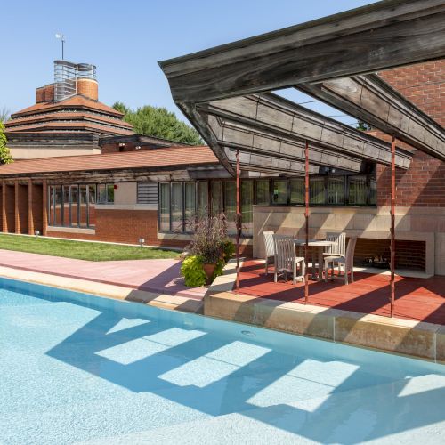A modern building with a slanted roof, next to a clear blue swimming pool, surrounded by trees and greenery, under a clear sky.