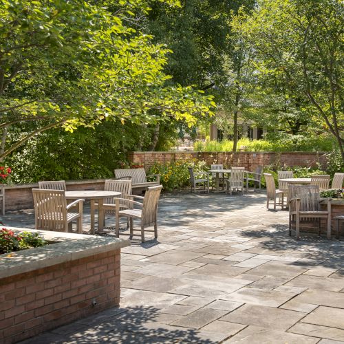 A serene garden patio with wooden tables and chairs, lush greenery, and stone paving, offering a peaceful outdoor setting.