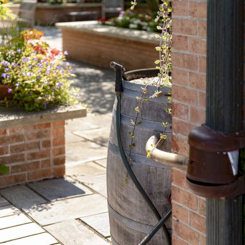 A garden scene with a wooden rain barrel, hose, and brick walls. Flower pots are visible in the background, suggesting a well-kept garden.