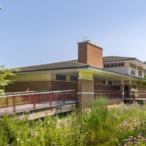 The image shows a modern house with a brick chimney, surrounded by greenery and flowers, set under a clear blue sky.