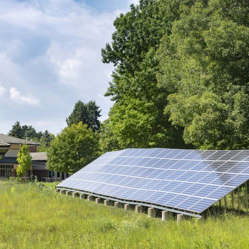 A solar panel array installed in a grassy area near a building, bordered by lush green trees under a partly cloudy sky.