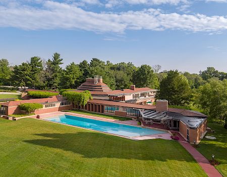 A large, architecturally distinct house with a swimming pool, surrounded by lush greenery and trees under a clear blue sky.