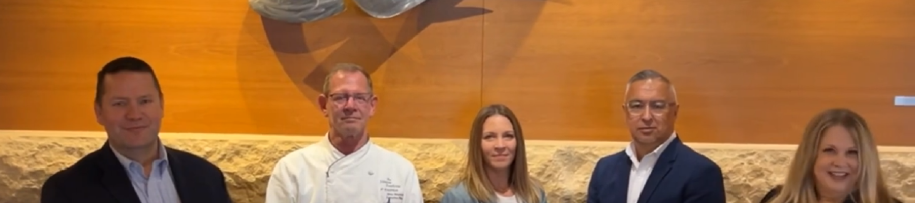 Five people standing in front of a wooden wall with a metal sculpture, smiling, dressed in formal attire and a chef's coat.