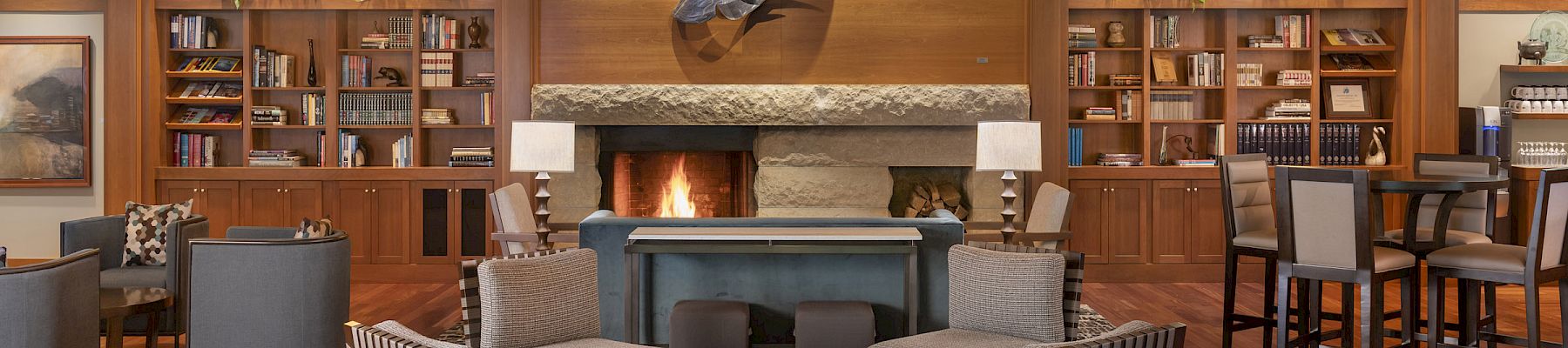 A cozy lounge with a fireplace, armchairs, bookshelves, and tables. The wooden ceiling and decor create a warm, inviting atmosphere.