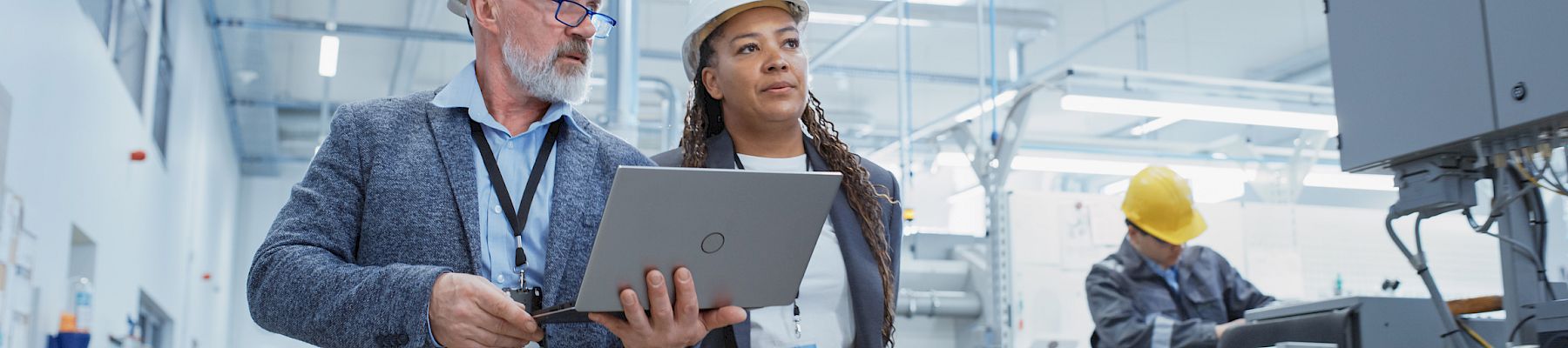 Two people in hard hats, one holding a laptop, walking through an industrial setting. Another person is working on machinery in the background.