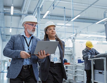 Two people in hard hats, one holding a laptop, walking through an industrial setting. Another person is working on machinery in the background.