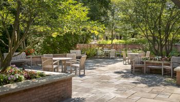 A peaceful outdoor patio with wooden furniture, surrounded by trees and greenery. Flowers adorn the brick planters, creating a cozy setting.