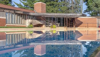 A modern house with large windows and a pool reflecting the building, surrounded by trees under a clear sky.