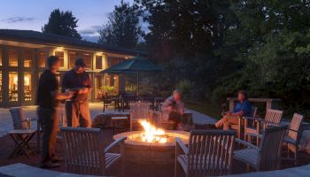 People gather around a fire pit in a cozy outdoor setting with chairs, near a building with lit windows, under a twilight sky.