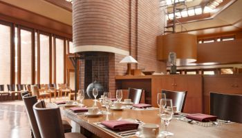 A modern dining room with a long table, set with glasses, plates, and napkins, featuring natural light and brick walls.