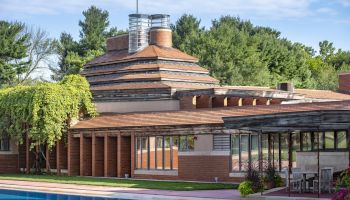 The image shows a modern architectural building with a unique pyramidal roof, surrounded by greenery and a reflecting pool in front.