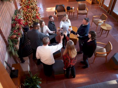 A group of people is gathered indoors near a decorated Christmas tree, raising glasses in a toast.