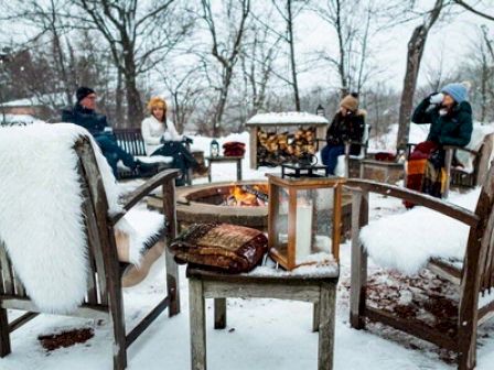 People are sitting around a fire pit in snowy surroundings, with fluffy blankets draped over chairs.