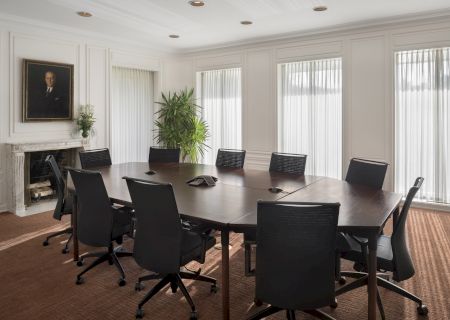 A conference room with a large oval table, black chairs, a fireplace with a portrait, and potted plants by tall windows.
