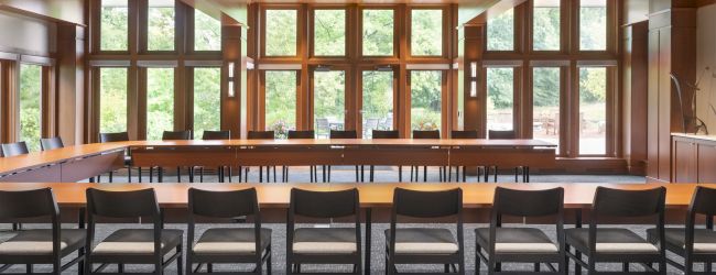 A spacious conference room with wooden beams, large windows, and numerous chairs arranged around a long rectangular table.