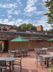 An outdoor patio with tables, chairs, and green umbrellas, surrounded by flowers, near a building with a wooden and brick facade.