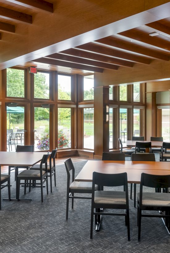 A modern, well-lit meeting room with wooden tables, black chairs, and large windows showing a green outdoor view. A screen is mounted on the wall.