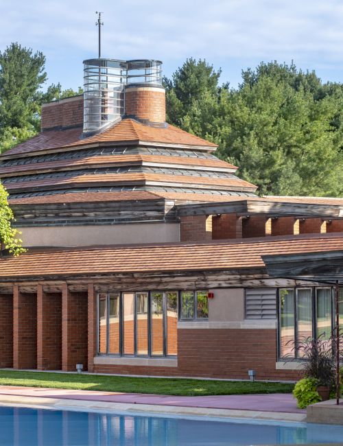 The image shows a uniquely designed building with a circular tower, surrounded by trees and featuring a nearby pool.