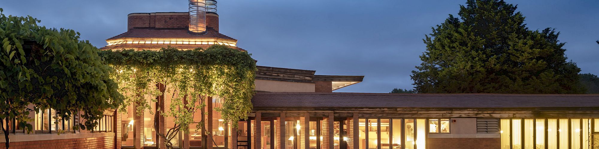 A modern building with large windows and a pool, surrounded by trees and evening sky, reflecting lights on the water's surface.