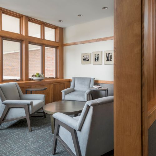 A small waiting area with gray chairs, a round table, wall art, large windows, and a quirky owl statue holding a book by the corner window.