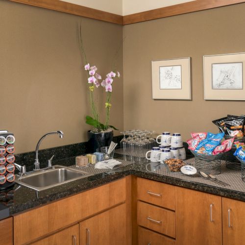 A kitchenette with coffee maker, snacks, drinks, a sink, glasses, and orchids on the counter next to framed wall art.