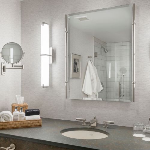 A modern bathroom featuring a large mirror, vanity with toiletries, wall lights, and a chromed faucet, with a glimpse of a shower in the reflection.