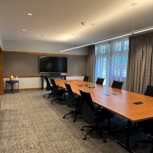 A modern conference room with a long table, chairs, a TV on the wall, and windows with curtains.