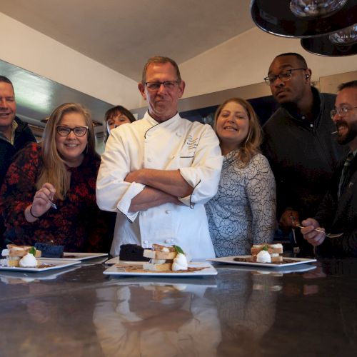 A chef stands proudly at a table with six people eagerly observing plated dishes in a kitchen setting.