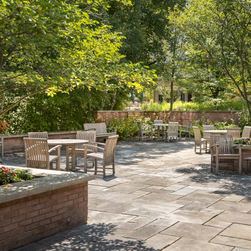 A serene garden patio with wooden tables and chairs, surrounded by greenery and flower beds, creating a peaceful outdoor seating area.