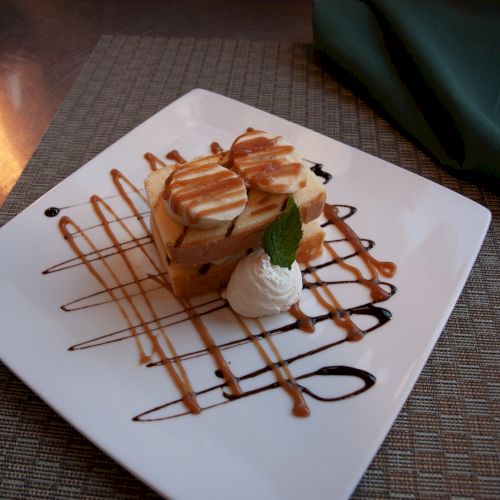 A dessert on a white plate with caramel and chocolate drizzle, topped with whipped cream and mint, is beautifully presented on a table.