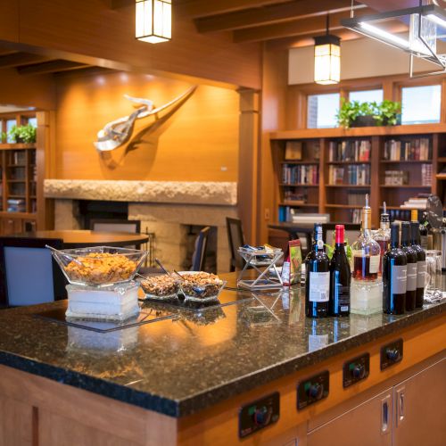 A cozy room with a kitchen island, snacks, and wine bottles, surrounded by a library area with bookshelves and seating spaces.
