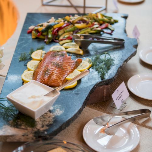 A platter with smoked salmon, lemon slices, grilled vegetables, and a dip on a stone serving tray, accompanied by plates and crackers.