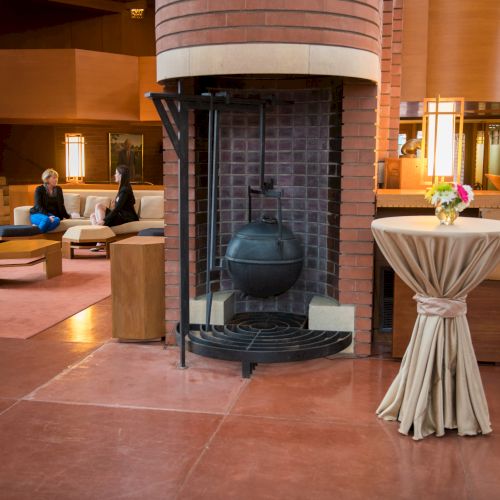 A modern interior features a central fireplace, a table with a tablecloth and flowers, and people seated on a couch in the background.
