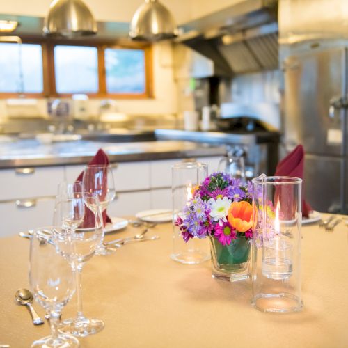 A table set with glassware and napkins, centered with a flower arrangement, in a kitchen with stainless steel appliances in the background.