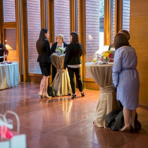 People are gathered around cocktail tables in a warmly lit room with large windows and a buffet in the background.
