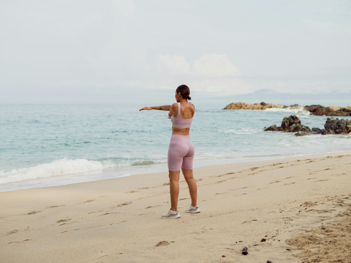 A person in athletic wear exercising on a sandy beach, facing the ocean, with rocks and calm waves in the background.