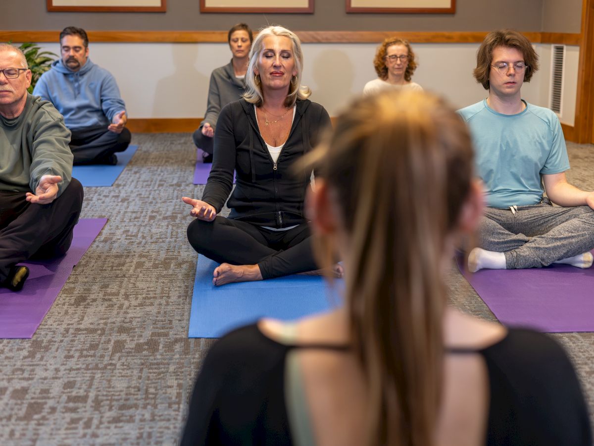 A group of adults sits on colorful mats in a circle, eyes closed or focused, meditating as a facilitator leads from the foreground.