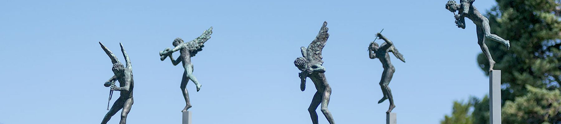 Sculptures of angels playing instruments are perched on tall poles against a backdrop of trees and a building with a red roof.