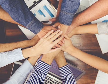 A diverse group forms a united teamwork circle, hands stacked together over a table with laptops and papers, signaling collaboration and trust.