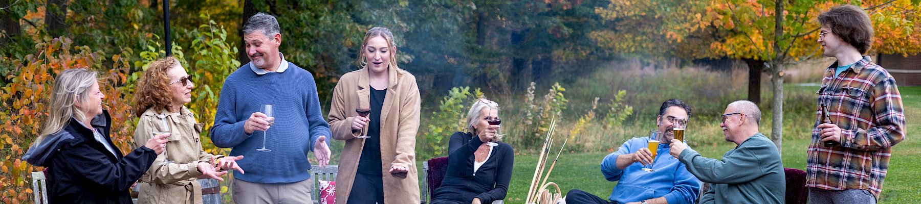 A group of friends sits and stands around a campfire in a park, chatting and toasting marshmallows on a cool autumn evening.