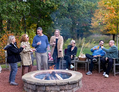 A group of friends sits and stands around a campfire in a park, chatting and toasting marshmallows on a cool autumn evening.