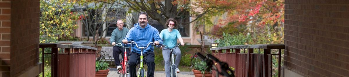 Three people riding bicycles on a covered path, with autumn trees in the background. The scene shows a group cycling toward the camera, framed by brick walls and a foreground bicycle rack.