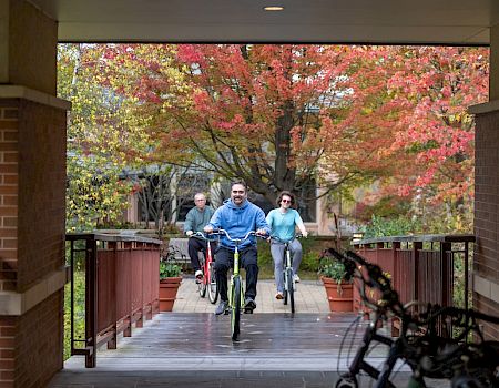 Three people riding bicycles on a covered path, with autumn trees in the background. The scene shows a group cycling toward the camera, framed by brick walls and a foreground bicycle rack.