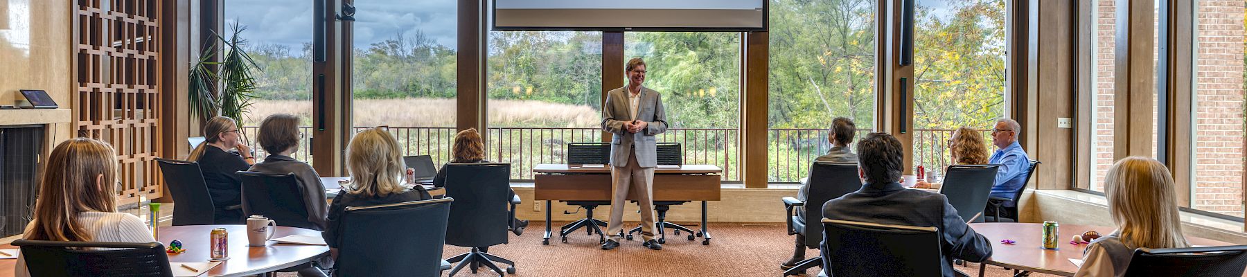 A presenter talks to a small audience in a bright conference room with a screen showing “Wingcorp” and a scenic outdoor view in the background.