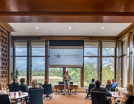 A presenter talks to a small audience in a bright conference room with a screen showing “Wingcorp” and a scenic outdoor view in the background.