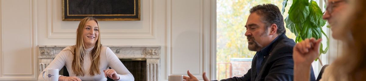 A business meeting in an office: a smiling woman at a table with a laptop while colleagues talk and coffee cups sit nearby.