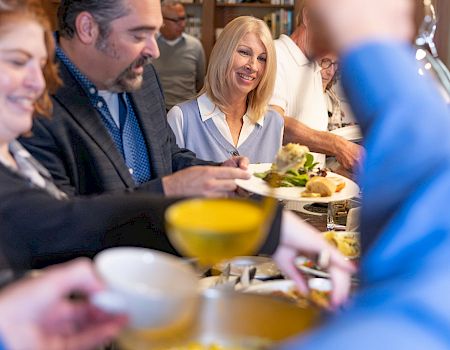 A group of coworkers in an office cafeteria, with a smiling woman serving food on plates during a meal.