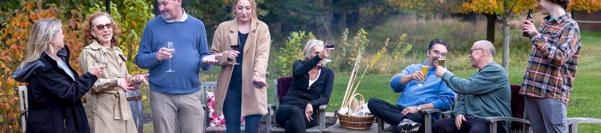 A group of friends gathers outdoors around a stone fire pit, toasting with drinks on a fall evening.